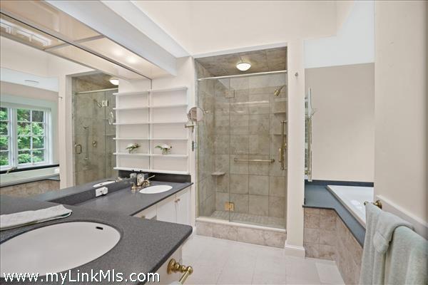 The spacious primary bathroom serves as a serene retreat, featuring a double vanity, a large glass-enclosed shower, a deep soaking tub, and custom floor-to-ceiling built-in shelving for linen and decor storage