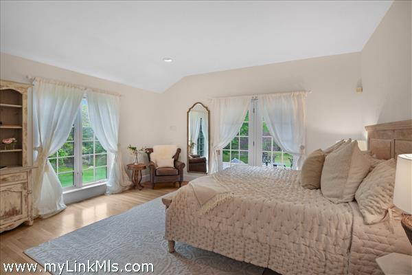 The sun-drenched primary bedroom serves as a private sanctuary, featuring expansive windows, elegant drapery, and French doors that open directly to a tranquil outdoor deck